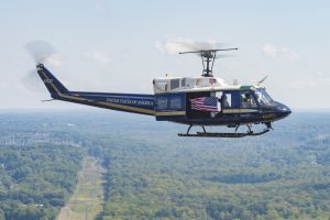 A blue and white UH-1N Iroquois Huey-style helicopter flies an American Flag in 2015 during a military event. Credit: U.S. Air Force photo/Airman 1st Class Ryan J. Sonnier